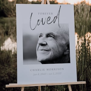 May include: A memorial sign on a wooden easel. The sign reads "Forever Loved" above a black and white photo of a smiling man. Below the photo is the name "Charlie Morrison" and dates "Jan 8, 1940 - Oct 1, 2023".