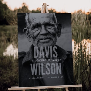 May include: A black and white memorial sign on a wooden easel. The sign features a portrait of an older man and the text "DAVIS IN LOVING MEMORY WILSON FEBRUARY 1994 - DECEMBER 2024". The background is a natural outdoor scene.
