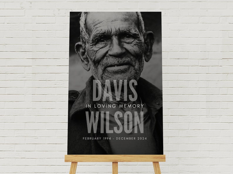 May include: A black and white memorial sign featuring a portrait of an older man. The sign reads "DAVIS IN LOVING MEMORY WILSON" with dates "FEBRUARY 1994 - DECEMBER 2024". The sign is displayed on a wooden easel.