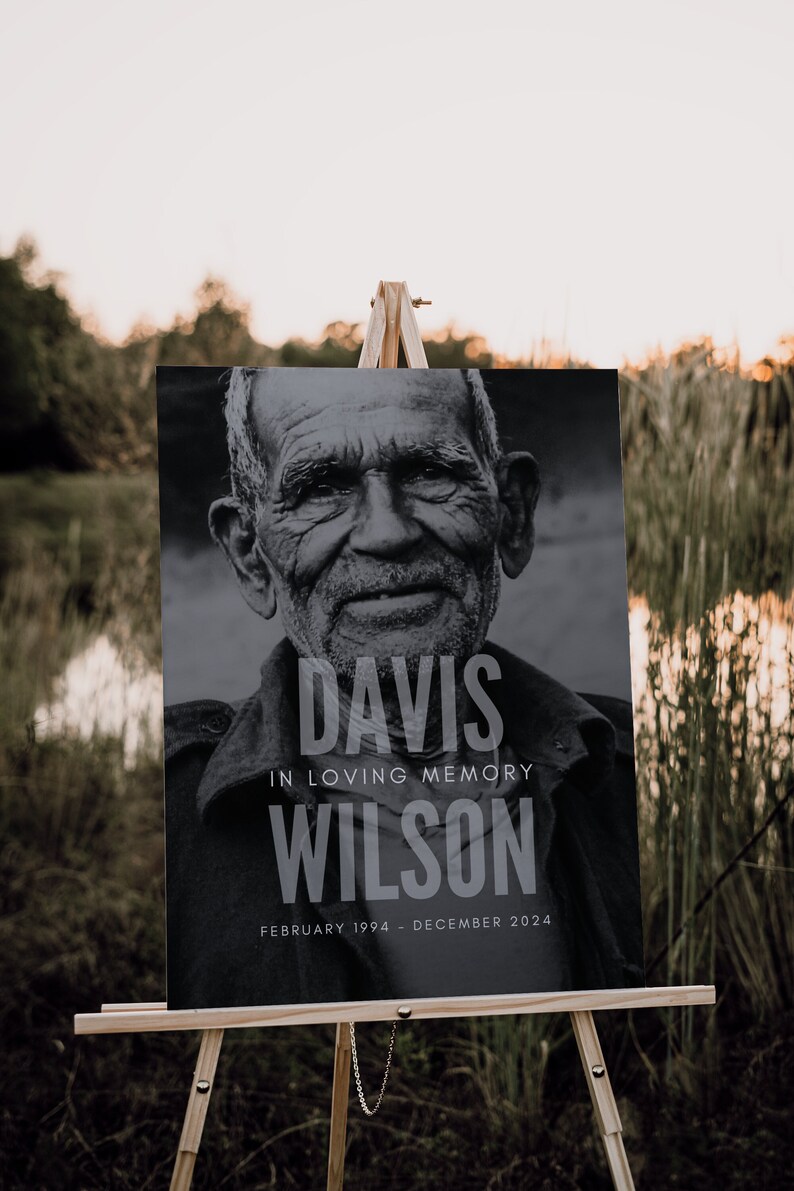 May include: A black and white memorial sign on a wooden easel. The sign features a portrait of an older man and the text "DAVIS IN LOVING MEMORY WILSON FEBRUARY 1994 - DECEMBER 2024". The background is a natural outdoor scene.