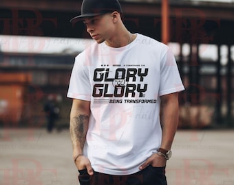 May include: White t-shirt with the text "GLORY TO GLORY BEING TRANSFORMED" in black. The shirt has a crew neck and short sleeves. The text is in a bold, stylized font. The shirt is worn by a person.