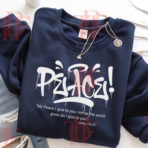 May include: Navy blue sweatshirt with the word "Peace" in white graffiti-style lettering. The sweatshirt also features the Bible verse John 14:27. A silver necklace with a small pendant rests on the sweatshirt.