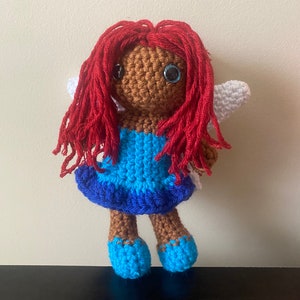 May include: A crocheted doll with brown skin, red hair, blue eyes, and a blue and white dress. The doll is wearing blue shoes and has white wings.