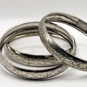May include: Three silver-colored bangles with floral and leaf patterns. The bangles are hinged and appear to be made of metal. The intricate designs are visible on the surface of the bangles, which are arranged in a slightly overlapping manner.