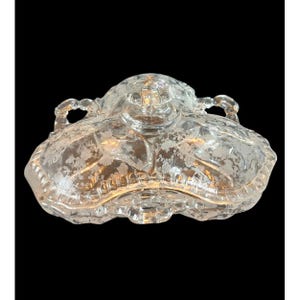 May include: Clear glass serving dish with a lid. The dish has a floral pattern etched into the glass. It features two handles and a decorative knob on the lid. The dish is likely used for serving food or as a decorative piece.