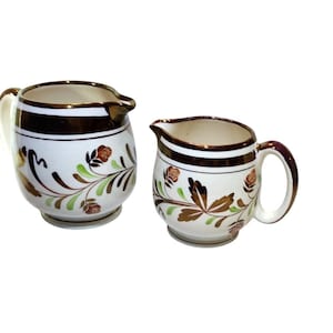 May include: Two ceramic cream pitchers with a white base, brown trim, and floral designs in brown, green, and red. The pitchers have a handle and a spout for pouring. The design is vintage.