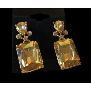May include: A pair of gold-toned drop earrings. Each earring features a large, rectangular, amber-colored gemstone, accented with smaller clear crystals and a teardrop-shaped stone at the top. The earrings are set against a black background.