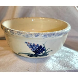 Può includere: Ciotola in ceramica color crema con un motivo floreale blu. La ciotola ha un bordo maculato blu e un fiore blu dipinto a mano con foglie verdi. Un oggetto decorativo per la cucina.