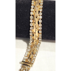 May include: A gold-toned bracelet featuring a central row of elongated, polished gold links flanked by two parallel rows of clear, sparkling round rhinestones. The piece includes a secure metal clasp at the end, showcasing a multi-strand design.