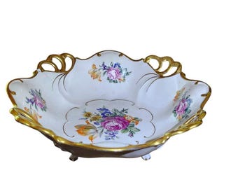 Könnte beinhalten: Eine weiße Porzellan-Servierschale mit gewelltem Rand und goldfarbenem Rand. Die Schale hat ein Blumendesign mit rosa, lila, blauen und gelben Blumen. Die Schale hat drei kleine Füße und dekorative Griffe.