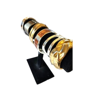 May include: A collection of bracelets displayed on a black velvet stand. The bracelets include gold, silver, and wood-tone designs. The stand is rectangular with a vertical support, showcasing the jewelry against a white background.