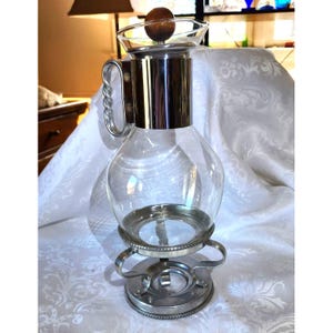 May include: A clear glass coffee carafe with a silver metal base and accents. The carafe has a bulbous body, a silver metal collar, and a glass top with a wooden knob. The handle is silver metal. The carafe sits on a silver metal stand.