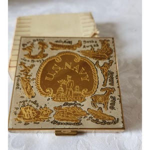 May include: A vintage compact mirror with a gold-colored design featuring the words "U.S. NAVY" in a central circle. The design includes illustrations of ships, buildings, and figures, with the word "Annapolis" visible.