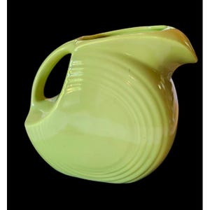 May include: A vintage, chartreuse-colored pitcher with a curved handle and a spout. The pitcher has a rounded body with concentric, ridged detailing. The pitcher is made of ceramic and is set against a black background.
