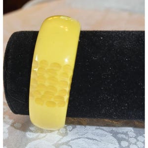 May include: A pale yellow bangle bracelet with a textured pattern on one side. The bracelet is displayed on a black velvet surface, showcasing its smooth, rounded shape and color.