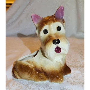 May include: A ceramic figurine of a terrier dog with a hollow back, featuring a cream-colored face and chest, brown fur on the body and ears, black eyes, a black nose, and a small open mouth with a dark pink tongue visible inside the mouth.