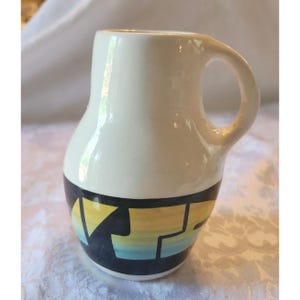 May include: A cream-colored ceramic pitcher with a single handle. The lower portion features a horizontal band with a black geometric design containing a gradient of yellow, orange, and light blue horizontal stripes within the shapes.