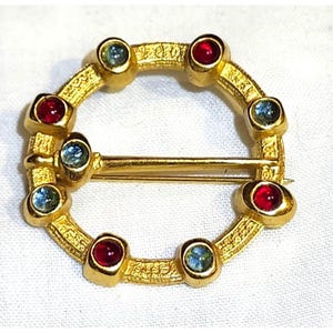 May include: A circular gold-tone brooch featuring a textured surface and a pin mechanism across the center. The frame is set with alternating round cabochon stones in deep red and light blue, creating a symmetrical pattern around the circular band.