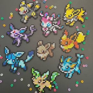 May include: Nine different Pokemon characters made with colorful perler beads. The characters include Eevee, Flareon, Jolteon, Vaporeon, Espeon, Umbreon, Leafeon, Glaceon, and Sylveon. The beads are arranged in a circle on a black background. The text '@Qtsliebeadz' is in the bottom right corner of the image.