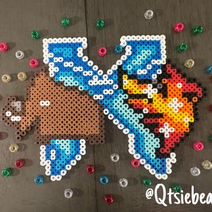 May include: A brown bear with blue wings and a red and yellow fire design. The bear is made of perler beads and is a pixel art design. The image includes the text "@Qtsicbeadz".