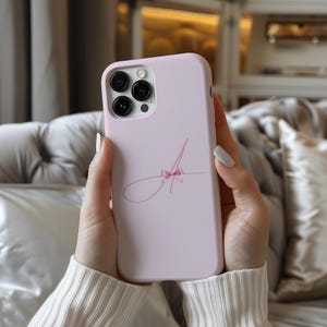 May include: A pink phone case with a black camera lens and a pink ribbon design. The case has a white letter "A" with a pink ribbon on it.