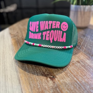 May include: Green trucker hat with a pink glitter design that says "Save Water Drink Tequila" and a pink smiley face. The hat has a white and green beaded band with the words "Mama  ♥  MegZ".