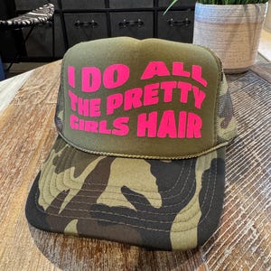 May include: An olive green trucker hat with a camouflage brim. The hat displays the text "I DO ALL THE PRETTY GIRLS HAIR" in pink. It has a mesh back and a curved brim. The hat is a stylish accessory.