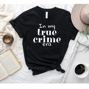 May include: Black t-shirt with the white text "In my true crime era." The shirt is styled with a knot at the bottom. A hat, coffee, and a book are in the background.