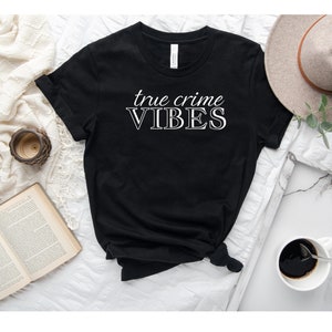 May include: Black t-shirt with white text that reads "true crime VIBES". The shirt is laid flat on a white surface with a book, a clock, a cup, and a hat in the background.