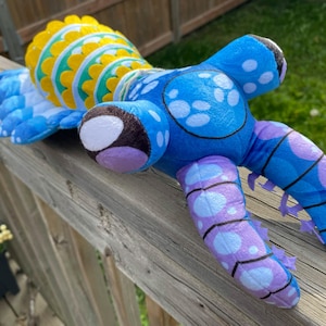 May include: A plush toy featuring a bright yellow and green patterned head with scalloped edges. The body is blue with purple and light blue circular spots, featuring segmented limbs and dark brown accents on the feet, crafted from soft fabric.