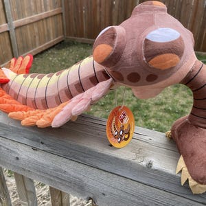 May include: A plush toy shaped like an Anomalocaris, featuring a brown, tan, and orange segmented body with large, dark brown eyes. The creature has orange fins along its sides and a pair of brown, claw-like appendages at the front with a round tag.