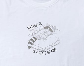 Op de afbeelding: Wit T-shirt met een zwart-witte illustratie van een wasbeer die in een kussen slaapt. De tekst "Sleeping in is a state of mind" staat onder de illustratie.