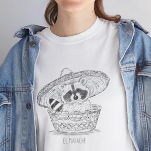 Może przedstawiać: Biała koszulka z czarno-białą ilustracją szopa pracza w sombrero siedzącego w wiklinowym koszu. Pod spodem nadrukowano napis "EL MAPACHE".