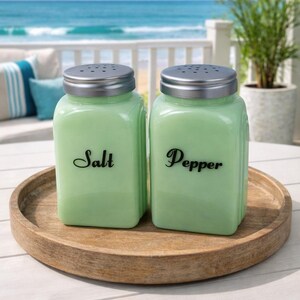 May include: Two light green glass salt and pepper shakers with silver lids sit on a wooden tray. The word "Salt" is printed on one shaker, and "Pepper" on the other. The background shows a beach scene.