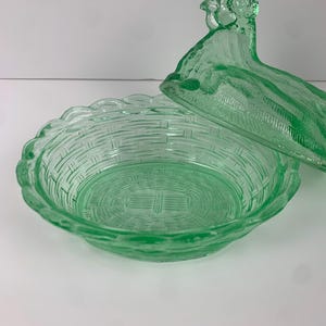 May include: A green glass basket-shaped dish with a lid. The dish has a woven basket design and a scalloped edge. The lid is shaped like a bird. The glass is a light green color.