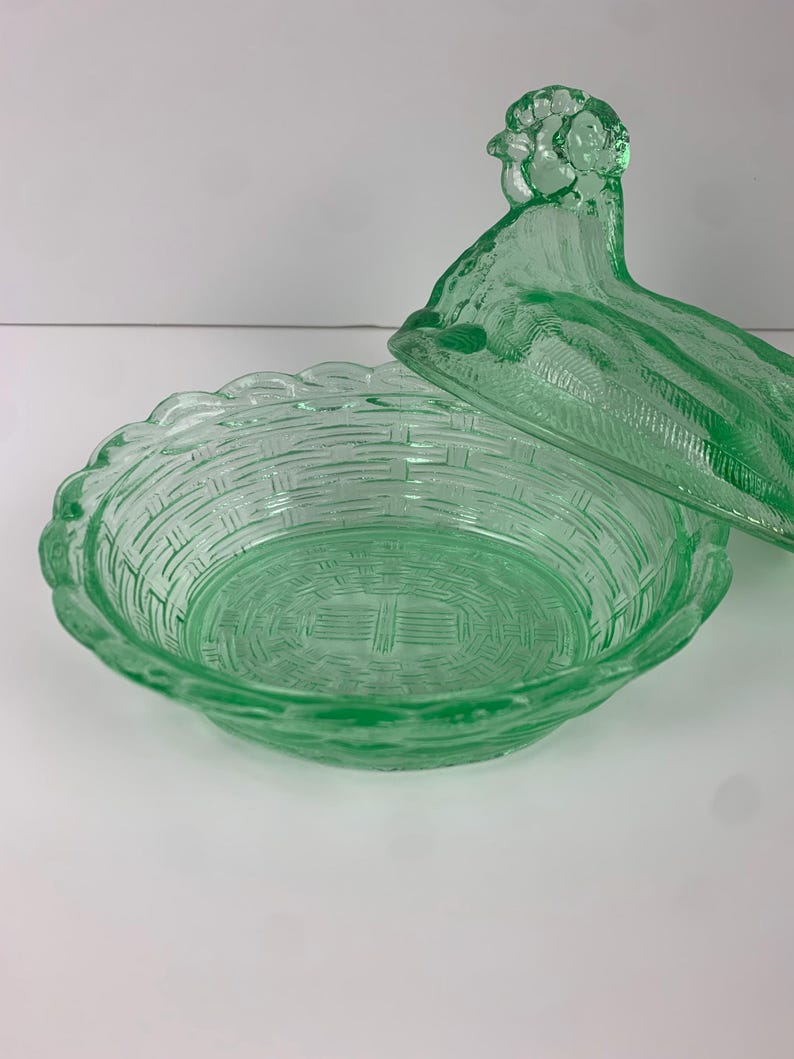 May include: A green glass basket-shaped dish with a lid. The dish has a woven basket design and a scalloped edge. The lid is shaped like a bird. The glass is a light green color.