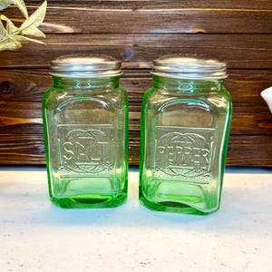May include: Two vintage green glass salt and pepper shakers with silver lids. The salt shaker is labeled "SALT" and the pepper shaker is labeled "PEPPER". The shakers are rectangular with rounded corners.