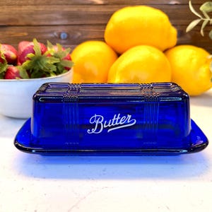 Può includere: Un portaburro rettangolare in vetro blu cobalto con un coperchio caratterizzato da un motivo a griglia e la parola Butter stampata in corsivo bianco sul davanti. Il piatto ha una base liscia e un coperchio strutturato con linee in rilievo.