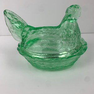 May include: A green glass hen-shaped covered dish. The hen is perched atop a nest-shaped base. The glass has a textured surface, and the dish is a vibrant shade of green. This decorative piece is likely vintage.