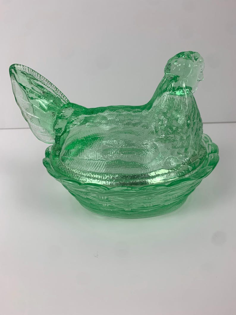 May include: A green glass hen-shaped covered dish. The hen is perched atop a nest-shaped base. The glass has a textured surface, and the dish is a vibrant shade of green. This decorative piece is likely vintage.