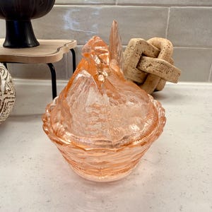 May include: A vintage, peach-colored glass hen-on-nest candy dish. The dish features a textured, ruffled design around the base and a detailed hen lid. This decorative piece is a charming addition to any kitchen or home decor.