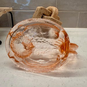 May include: A clear, pink-tinted glass dish with a textured surface and a decorative rim. The dish has a circular shape with a small handle. A light-colored, abstract sculpture is in the background.