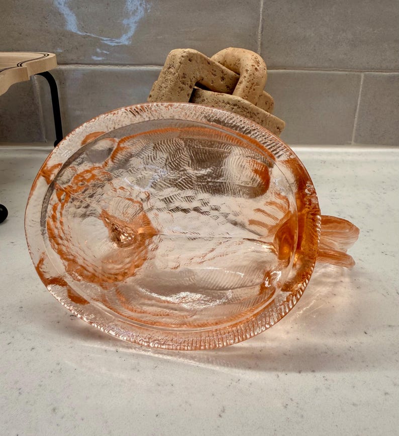 May include: A clear, pink-tinted glass dish with a textured surface and a decorative rim. The dish has a circular shape with a small handle. A light-colored, abstract sculpture is in the background.