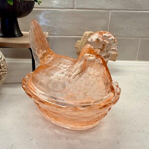 May include: A pink glass hen-shaped covered dish. The hen is sitting in a basket-like base. The glass has a textured surface. The dish is likely used for serving or storing food.