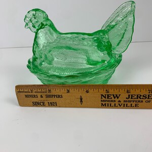 May include: A green glass hen-shaped dish with a textured surface, sitting on a woven-style base. The hen has detailed features, including a comb and tail feathers. A wooden ruler is in the foreground.