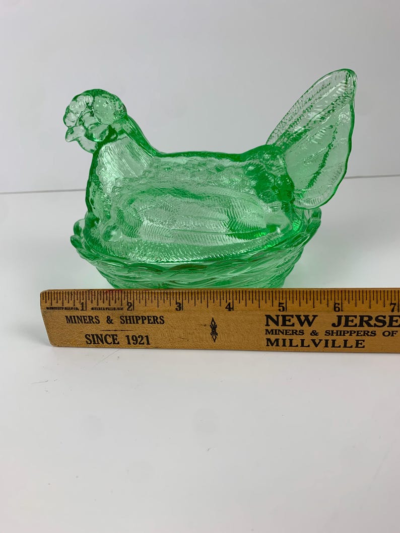 May include: A green glass hen-shaped dish with a textured surface, sitting on a woven-style base. The hen has detailed features, including a comb and tail feathers. A wooden ruler is in the foreground.