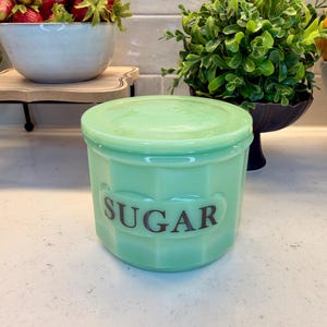 Puede incluir: Un tarro de azúcar de cristal verde claro con tapa a juego. El recipiente cilíndrico presenta un diseño exterior geométrico facetado y la palabra SUGAR impresa en letras mayúsculas de color marrón oscuro en el centro frontal.