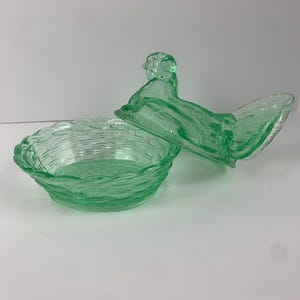 May include: A green glass hen-shaped covered dish. The dish has a basket weave design and the lid is shaped like a hen. The dish is made of a light green glass.