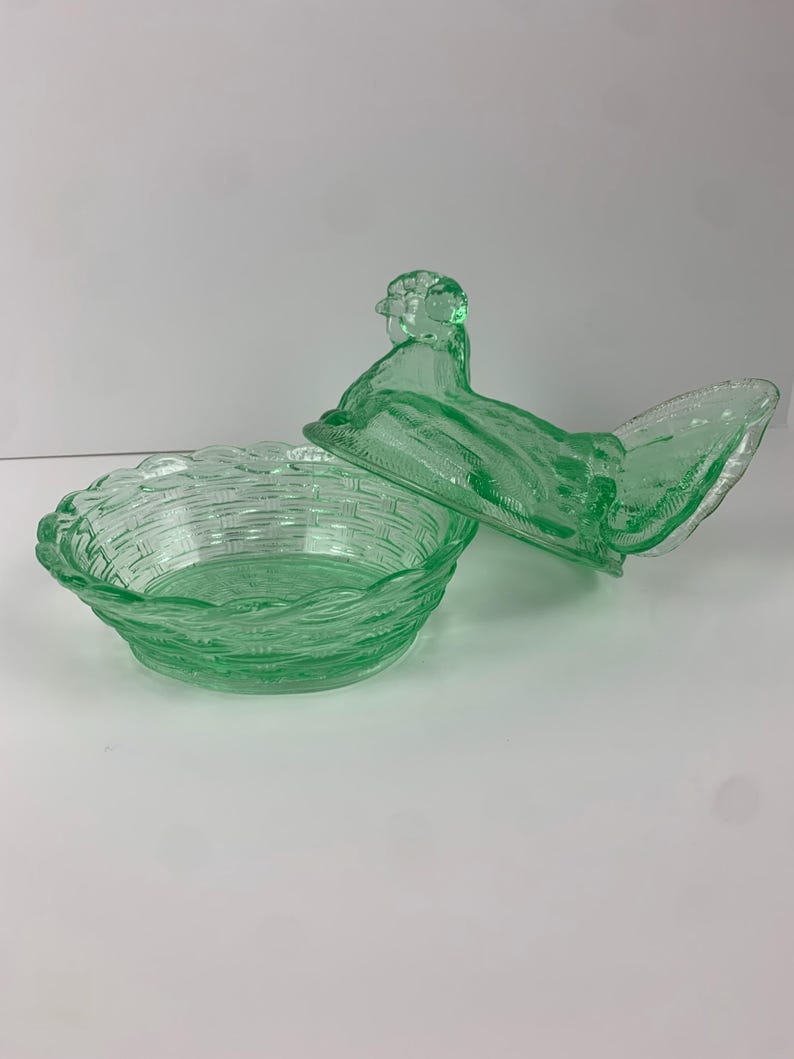 May include: A green glass hen-shaped covered dish. The dish has a basket weave design and the lid is shaped like a hen. The dish is made of a light green glass.