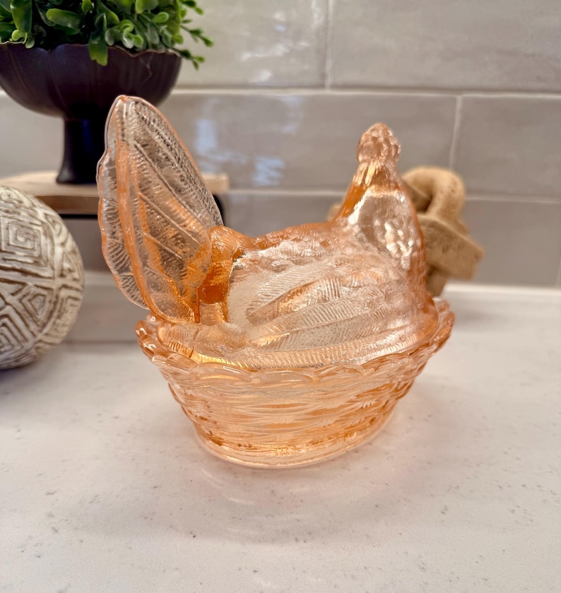 May include: A decorative, peach-colored glass hen-shaped dish. The hen is perched on a woven basket-like base, with detailed wing and feather textures. The dish is likely used for serving or display.